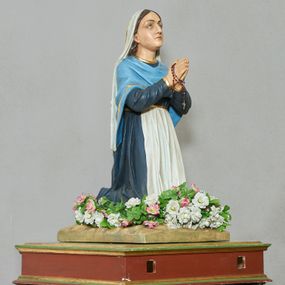 Zdjęcie nr 1: Rzeźba pełna, na podstawie imitującej skałę. Zamontowana na wysokiej podstawie na planie zbliżonym do kwadratu, o krawędziach ujętych profilowanymi listewkami, z dwoma otworami na drążki. Bernadeta ukazana w pozycji klęczącej, z rękami złożonymi w geście modlitwy, z lekko uniesioną głową. Twarz ma pociągłą, o migdałowatych oczach skierowanych w górę, dużym nosie i małych ustach; okoloną długimi brązowymi włosami opadającymi na plecy. Ubrana jest w granatową suknię, o złoconych lamówkach, przepasaną w talii białym fartuchem, na ramiona i plecy ma narzuconą niebieską chustę o złoconych lamówkach, na głowie ma biały welon. Polichromia ciała naturalistyczna. Wokół nóg figury zamocowane sztuczne kwiaty.



