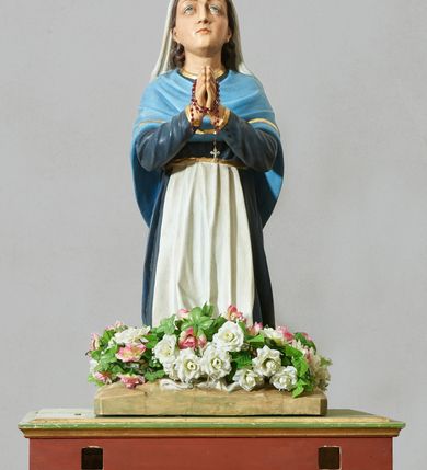 Zdjęcie nr 2: Rzeźba pełna, na podstawie imitującej skałę. Zamontowana na wysokiej podstawie na planie zbliżonym do kwadratu, o krawędziach ujętych profilowanymi listewkami, z dwoma otworami na drążki. Bernadeta ukazana w pozycji klęczącej, z rękami złożonymi w geście modlitwy, z lekko uniesioną głową. Twarz ma pociągłą, o migdałowatych oczach skierowanych w górę, dużym nosie i małych ustach; okoloną długimi brązowymi włosami opadającymi na plecy. Ubrana jest w granatową suknię, o złoconych lamówkach, przepasaną w talii białym fartuchem, na ramiona i plecy ma narzuconą niebieską chustę o złoconych lamówkach, na głowie ma biały welon. Polichromia ciała naturalistyczna. Wokół nóg figury zamocowane sztuczne kwiaty.



