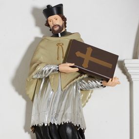 Zdjęcie nr 1: Rzeźba pełnoplastyczna, ustawiona na niskiej, nieregularnej podstawie i niewielkim prostopadłościennym postumencie umieszczonym na konsoli. Figura św. Jana Nepomucena jest zwrócona frontalnie, z głową przechyloną w prawo, w mocnym kontrapoście, o esowato wygiętej sylwetce. Święty w obu rękach zwróconych w lewo podtrzymuje księgę. Twarz ma okrągłą, o delikatnie zarysowanych oczach, szerokim nosie i otwartych ustach, okoloną brodą i krótkimi, kręconymi włosami. Święty ubrany jest w strój kanonika: czarną sutannę, posrebrzoną komżę oraz beżową almucję, na głowie ma czarny biret. Polichromia ciała naturalistyczna. 