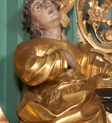 Zdjęcie nr 3: Rzeźba drążona z tyłu, ukazana frontalnie, w wykroku, z głową przechyloną delikatnie w tył, skierowaną w lewo. Prawa ręka ugięta, dłonią wskazuje na pierś, lewą święty przytrzymuje na biodrze zamkniętą księgę. Twarz ma podłużną, pełną, o młodzieńczych rysach, długim nosie, głęboko osadzonych i otwartych oczach zwróconych ku górze oraz delikatnie rozchylonych ustach. Włosy w postaci loków opadają na plecy. Święty ubrany w ściśle przylegającą do ciała tunikę przewiązaną w talii oraz otaczający postać płaszcz zawieszony na lewym ramieniu. Tunika delikatnie marszczona, ściśle przylegająca do ciała, zaś draperia płaszcza głęboko rzeźbiona, ekspresyjnie fałdowana. Przy lewej nodze postaci figura orła. Polichromia naturalistyczna w partiach ciała, szaty pozłacane, orzeł posrebrzany.