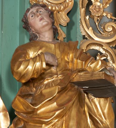 Zdjęcie nr 2: Rzeźba drążona z tyłu, ukazana frontalnie, w wykroku, z głową przechyloną delikatnie w tył, skierowaną w lewo. Prawa ręka ugięta, dłonią wskazuje na pierś, lewą święty przytrzymuje na biodrze zamkniętą księgę. Twarz ma podłużną, pełną, o młodzieńczych rysach, długim nosie, głęboko osadzonych i otwartych oczach zwróconych ku górze oraz delikatnie rozchylonych ustach. Włosy w postaci loków opadają na plecy. Święty ubrany w ściśle przylegającą do ciała tunikę przewiązaną w talii oraz otaczający postać płaszcz zawieszony na lewym ramieniu. Tunika delikatnie marszczona, ściśle przylegająca do ciała, zaś draperia płaszcza głęboko rzeźbiona, ekspresyjnie fałdowana. Przy lewej nodze postaci figura orła. Polichromia naturalistyczna w partiach ciała, szaty pozłacane, orzeł posrebrzany.