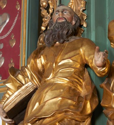 Zdjęcie nr 2: Rzeźba drążona z tyłu, zwrócona frontalnie, z głową skierowaną ku górze, o esowato wygiętej sylwetce, w kontrapoście, z lewą nogą opartą na skale. Lewa ręka jest ugięta i uniesiona, prawą święty przytrzymuje księgę, opierając ją na biodrze. Twarz ma pociągłą, o starczych rysach, długim nosie, delikatnie rozchylonych ustach i oczach zwróconych w górę. Okolona jest długą brodą w formie opadających na pierś pukli. Włosy ma krótkie. Postać ubrana jest w przylegającą do ciała tunikę, delikatnie marszczoną w partii torsu i obficie fałdowaną u spodu oraz przewiązany diagonalnie płaszcz, zawieszony na prawym ramieniu, opadający na lewe biodro w postaci silnie giętej i obficie marszczonej tkaniny. Przy prawej stopie postaci głowa wołu. Polichromia naturalistyczna w partiach ciała i atrybutów, szaty pozłacane.
