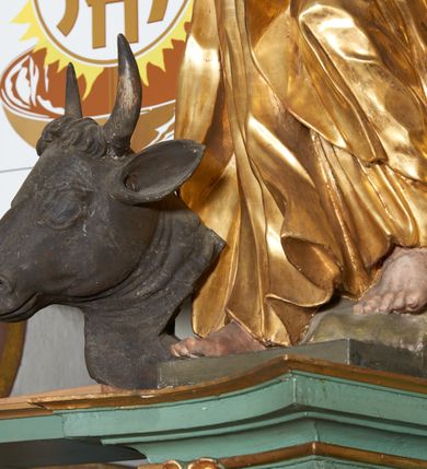 Zdjęcie nr 3: Rzeźba drążona z tyłu, zwrócona frontalnie, z głową skierowaną ku górze, o esowato wygiętej sylwetce, w kontrapoście, z lewą nogą opartą na skale. Lewa ręka jest ugięta i uniesiona, prawą święty przytrzymuje księgę, opierając ją na biodrze. Twarz ma pociągłą, o starczych rysach, długim nosie, delikatnie rozchylonych ustach i oczach zwróconych w górę. Okolona jest długą brodą w formie opadających na pierś pukli. Włosy ma krótkie. Postać ubrana jest w przylegającą do ciała tunikę, delikatnie marszczoną w partii torsu i obficie fałdowaną u spodu oraz przewiązany diagonalnie płaszcz, zawieszony na prawym ramieniu, opadający na lewe biodro w postaci silnie giętej i obficie marszczonej tkaniny. Przy prawej stopie postaci głowa wołu. Polichromia naturalistyczna w partiach ciała i atrybutów, szaty pozłacane.