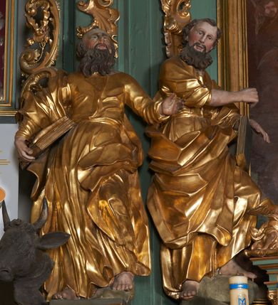 Zdjęcie nr 4: Rzeźba drążona z tyłu, zwrócona frontalnie, z głową skierowaną ku górze, o esowato wygiętej sylwetce, w kontrapoście, z lewą nogą opartą na skale. Lewa ręka jest ugięta i uniesiona, prawą święty przytrzymuje księgę, opierając ją na biodrze. Twarz ma pociągłą, o starczych rysach, długim nosie, delikatnie rozchylonych ustach i oczach zwróconych w górę. Okolona jest długą brodą w formie opadających na pierś pukli. Włosy ma krótkie. Postać ubrana jest w przylegającą do ciała tunikę, delikatnie marszczoną w partii torsu i obficie fałdowaną u spodu oraz przewiązany diagonalnie płaszcz, zawieszony na prawym ramieniu, opadający na lewe biodro w postaci silnie giętej i obficie marszczonej tkaniny. Przy prawej stopie postaci głowa wołu. Polichromia naturalistyczna w partiach ciała i atrybutów, szaty pozłacane.