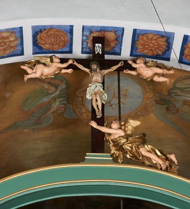 Zdjęcie nr 2: Belka tęczowa w formie łuku segmentowego nadwieszonego z krucyfiksem umieszczonym na środku, na profilowanym cokole. Do prostego krzyża przybita trzema gwoździami figura umarłego Chrystusa. Postać ukazana w lekkim zwisie, o szeroko rozłożonych ramionach, z głową opadającą na prawy bark, nogi lekko ugięte w kolanach. Twarz Chrystusa jest pociągła, szczupła, o głęboko osadzonych, zamkniętych oczach, długim, wąskim nosie i niewielkich ustach, okolona jest zarostem i długimi włosami w postaci wijących się pukli, opadających na plecy i ramiona. Na głowie korona cierniowa. Ciało o wyraźnie podkreślonej anatomii klatki piersiowej oraz muskulaturze ramion i nóg. Perizonium sięgające połowy ud, obficie fałdowane, zawiązane na lewym boku ze zwisem tkaniny. Ramiona poziomej belki krzyża podtrzymują małe, uskrzydlone aniołki, przepasane złotą tkaniną, zaś pionową, u spodu podtrzymuje nadlatujący anioł przedstawiony w pozycji półleżącej w silnie rozwianych i głęboko sfałdowanych szatach. Karnacja Chrystusa i aniołów malowana farbą olejną w kolorach naturalnych, perizonium białe, skrzydła i szaty aniołów pozłacane.