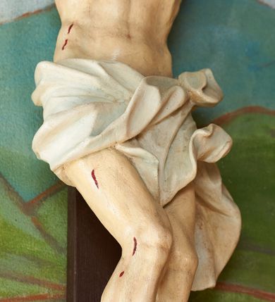 Zdjęcie nr 4: Figura umarłego Chrystusa przybita trzema gwoździami do krzyża o prostych ramionach. Ciało w lekkim zwisie, o szeroko rozłożonych ramionach, układających się w delikatny łuk, esowato wygiętej sylwetce, głowa opada bezwładnie na prawy bark, obie nogi ugięte w kolanach, stopy skrzyżowane w układzie prawa na lewą. Twarz trójkątna, szczupła, o zamkniętych oczach, dużym, prostym nosie i wąskich ustach, okolona krótką brodą i włosami silnie skręconymi w pukle. Na głowie szeroka, zielona korona cierniowa. Ciało wychudzone, o silnie podkreślonej muskulaturze oraz żebrach klatki piersiowej. Biodra przepasane białym, krótkim i silnie drapowanym perizonium, zawiązanym na lewym boku. Polichromia ciała naturalistyczna z zaznaczonymi śladami męki. Na zakończeniu pionowej belki krzyża banderola z napisem „I.N./R.I.”