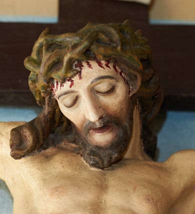 Zdjęcie nr 2: Figura umarłego Chrystusa przybita trzema gwoździami do krzyża o prostych ramionach. Ciało w lekkim zwisie, o szeroko rozłożonych ramionach, układających się w delikatny łuk, esowato wygiętej sylwetce, głowa opada bezwładnie na prawy bark, obie nogi ugięte w kolanach, stopy skrzyżowane w układzie prawa na lewą. Twarz trójkątna, szczupła, o zamkniętych oczach, dużym, prostym nosie i wąskich ustach, okolona krótką brodą i włosami silnie skręconymi w pukle. Na głowie szeroka, zielona korona cierniowa. Ciało wychudzone, o silnie podkreślonej muskulaturze oraz żebrach klatki piersiowej. Biodra przepasane białym, krótkim i silnie drapowanym perizonium, zawiązanym na lewym boku. Polichromia ciała naturalistyczna z zaznaczonymi śladami męki. Na zakończeniu pionowej belki krzyża banderola z napisem „I.N./R.I.”