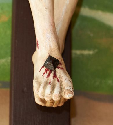 Zdjęcie nr 5: Figura umarłego Chrystusa przybita trzema gwoździami do krzyża o prostych ramionach. Ciało w lekkim zwisie, o szeroko rozłożonych ramionach, układających się w delikatny łuk, esowato wygiętej sylwetce, głowa opada bezwładnie na prawy bark, obie nogi ugięte w kolanach, stopy skrzyżowane w układzie prawa na lewą. Twarz trójkątna, szczupła, o zamkniętych oczach, dużym, prostym nosie i wąskich ustach, okolona krótką brodą i włosami silnie skręconymi w pukle. Na głowie szeroka, zielona korona cierniowa. Ciało wychudzone, o silnie podkreślonej muskulaturze oraz żebrach klatki piersiowej. Biodra przepasane białym, krótkim i silnie drapowanym perizonium, zawiązanym na lewym boku. Polichromia ciała naturalistyczna z zaznaczonymi śladami męki. Na zakończeniu pionowej belki krzyża banderola z napisem „I.N./R.I.”