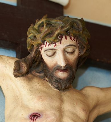 Zdjęcie nr 3: Figura umarłego Chrystusa przybita trzema gwoździami do krzyża o prostych ramionach. Ciało w lekkim zwisie, o szeroko rozłożonych ramionach, układających się w delikatny łuk, esowato wygiętej sylwetce, głowa opada bezwładnie na prawy bark, obie nogi ugięte w kolanach, stopy skrzyżowane w układzie prawa na lewą. Twarz trójkątna, szczupła, o zamkniętych oczach, dużym, prostym nosie i wąskich ustach, okolona krótką brodą i włosami silnie skręconymi w pukle. Na głowie szeroka, zielona korona cierniowa. Ciało wychudzone, o silnie podkreślonej muskulaturze oraz żebrach klatki piersiowej. Biodra przepasane białym, krótkim i silnie drapowanym perizonium, zawiązanym na lewym boku. Polichromia ciała naturalistyczna z zaznaczonymi śladami męki. Na zakończeniu pionowej belki krzyża banderola z napisem „I.N./R.I.”