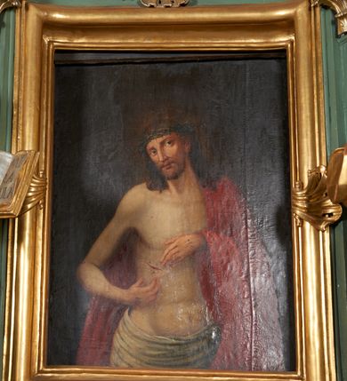 Zdjęcie nr 2: Obraz w formie stojącego prostokąta. Chrystus przedstawiony w półpostaci, zwrócony w trzech czwartych w lewo, z głową przechyloną na prawe ramię, obiema rękami wskazuje na ranę w prawym boku. Twarz podłużna, modelowana światłocieniem, o migdałowatych oczach, prostym nosie i pełnych ustach, okolona jasną, krótką brodą. Włosy brązowe, lekko kręcone, opadają na ramiona. Na głowie korona cierniowa. Chrystus w partii bioder przepasany białą, drapowaną tkaniną, na lewe ramię i plecy narzucony czerwony płaszcz. Ciało szczupłe, o miękkim modelunku światłocieniowym, delikatnie zarysowanym torsie, z zaznaczonymi śladami męki na dłoniach i prawym boku. Tło ciemnobrązowe, rozjaśnione wokół głowy postaci. Obraz utrzymany w ciepłej tonacji.