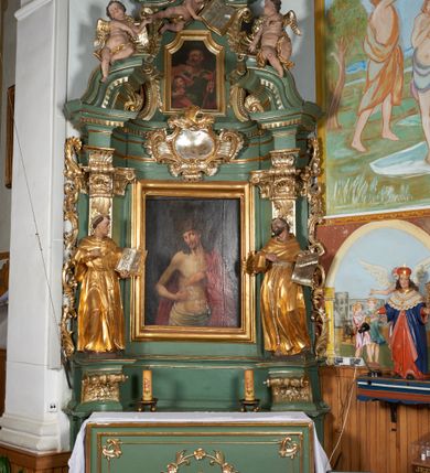 Zdjęcie nr 6: Obraz w formie stojącego prostokąta. Chrystus przedstawiony w półpostaci, zwrócony w trzech czwartych w lewo, z głową przechyloną na prawe ramię, obiema rękami wskazuje na ranę w prawym boku. Twarz podłużna, modelowana światłocieniem, o migdałowatych oczach, prostym nosie i pełnych ustach, okolona jasną, krótką brodą. Włosy brązowe, lekko kręcone, opadają na ramiona. Na głowie korona cierniowa. Chrystus w partii bioder przepasany białą, drapowaną tkaniną, na lewe ramię i plecy narzucony czerwony płaszcz. Ciało szczupłe, o miękkim modelunku światłocieniowym, delikatnie zarysowanym torsie, z zaznaczonymi śladami męki na dłoniach i prawym boku. Tło ciemnobrązowe, rozjaśnione wokół głowy postaci. Obraz utrzymany w ciepłej tonacji.