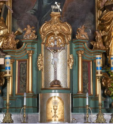 Zdjęcie nr 1: Tabernakulum w formie sześcianu, z wypukłym frontem. Drzwiczki w formie stojącego prostokąta zamkniętego łukiem półkolistym z aplikacją przedstawiającą kielich z hostią z monogramem IHS w glorii. Tron wystawienia architektoniczny, trójosiowy. Oś środkowa w postaci obrotowej niszy ujętej w profilowaną ramę, z konchą utworzoną z rocaille'u, zakończona ornamentalnym zwieńczeniem dekorowanym muszlą, małżowiną i kartuszem, które tworzą postument dla rzeźby baranka z krzyżem. Osie boczne na cokole, cofniętym pod kątem w tył, ujęte dwiema parami kolumn różnej wysokości o kapitelach kompozytowych, dźwigających pełne belkowanie w formie spływu, wyłamane w ich partii, na których znajdują się siedzące i klęczące figury aniołów. Między kolumnami prostokątne płyciny z wotami. Struktura polichromowana w kolorze jasnozielonym z pozłoconymi profilami i ornamentyką.