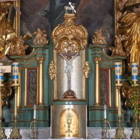 Zdjęcie nr 1: Tabernakulum w formie sześcianu, z wypukłym frontem. Drzwiczki w formie stojącego prostokąta zamkniętego łukiem półkolistym z aplikacją przedstawiającą kielich z hostią z monogramem IHS w glorii. Tron wystawienia architektoniczny, trójosiowy. Oś środkowa w postaci obrotowej niszy ujętej w profilowaną ramę, z konchą utworzoną z rocaille'u, zakończona ornamentalnym zwieńczeniem dekorowanym muszlą, małżowiną i kartuszem, które tworzą postument dla rzeźby baranka z krzyżem. Osie boczne na cokole, cofniętym pod kątem w tył, ujęte dwiema parami kolumn różnej wysokości o kapitelach kompozytowych, dźwigających pełne belkowanie w formie spływu, wyłamane w ich partii, na których znajdują się siedzące i klęczące figury aniołów. Między kolumnami prostokątne płyciny z wotami. Struktura polichromowana w kolorze jasnozielonym z pozłoconymi profilami i ornamentyką.