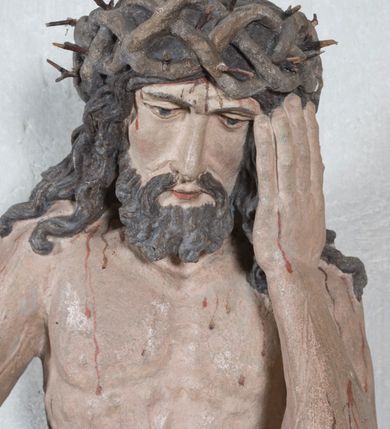 Zdjęcie nr 3: Figura pełnoplastyczna przedstawiająca Chrystusa Frasobliwego. Chrystus ukazany w pozycji siedzącej na kamiennej ławie, wspierający lewą ręką głowę, a prawą opierający na prawym kolanie, której dłoń ułożona jest do trzymania niezachowanego atrybutu. Ciało o silnie podkreślonej muskularnej i krępej budowie. Twarz podłużna, z długim i wąskim nosem, oczami skierowanymi w dół, okolona gęstą i kręconą brodą. Włosy długie, ciemnobrązowe, skręcone w pojedyncze pukle opadają na ramiona i plecy. Na głowie Chrystus ma założoną szeroką koronę cierniową, złożoną z przeplatających się symetrycznie gałązek z wystającymi cierniami. Perizonium białe, ciasno oplatające biodra, silnie drapowane. Polichromia w odsłoniętych partiach ciała naturalistyczna. Spod korony cierniowej oraz po całym ciele spływają drobne strużki krwi.

