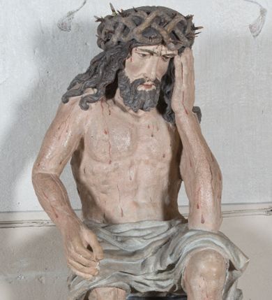 Zdjęcie nr 1: Figura pełnoplastyczna przedstawiająca Chrystusa Frasobliwego. Chrystus ukazany w pozycji siedzącej na kamiennej ławie, wspierający lewą ręką głowę, a prawą opierający na prawym kolanie, której dłoń ułożona jest do trzymania niezachowanego atrybutu. Ciało o silnie podkreślonej muskularnej i krępej budowie. Twarz podłużna, z długim i wąskim nosem, oczami skierowanymi w dół, okolona gęstą i kręconą brodą. Włosy długie, ciemnobrązowe, skręcone w pojedyncze pukle opadają na ramiona i plecy. Na głowie Chrystus ma założoną szeroką koronę cierniową, złożoną z przeplatających się symetrycznie gałązek z wystającymi cierniami. Perizonium białe, ciasno oplatające biodra, silnie drapowane. Polichromia w odsłoniętych partiach ciała naturalistyczna. Spod korony cierniowej oraz po całym ciele spływają drobne strużki krwi.

