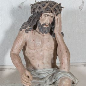 Zdjęcie nr 1: Figura pełnoplastyczna przedstawiająca Chrystusa Frasobliwego. Chrystus ukazany w pozycji siedzącej na kamiennej ławie, wspierający lewą ręką głowę, a prawą opierający na prawym kolanie, której dłoń ułożona jest do trzymania niezachowanego atrybutu. Ciało o silnie podkreślonej muskularnej i krępej budowie. Twarz podłużna, z długim i wąskim nosem, oczami skierowanymi w dół, okolona gęstą i kręconą brodą. Włosy długie, ciemnobrązowe, skręcone w pojedyncze pukle opadają na ramiona i plecy. Na głowie Chrystus ma założoną szeroką koronę cierniową, złożoną z przeplatających się symetrycznie gałązek z wystającymi cierniami. Perizonium białe, ciasno oplatające biodra, silnie drapowane. Polichromia w odsłoniętych partiach ciała naturalistyczna. Spod korony cierniowej oraz po całym ciele spływają drobne strużki krwi.

