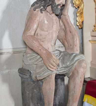Zdjęcie nr 2: Figura pełnoplastyczna przedstawiająca Chrystusa Frasobliwego. Chrystus ukazany w pozycji siedzącej na kamiennej ławie, wspierający lewą ręką głowę, a prawą opierający na prawym kolanie, której dłoń ułożona jest do trzymania niezachowanego atrybutu. Ciało o silnie podkreślonej muskularnej i krępej budowie. Twarz podłużna, z długim i wąskim nosem, oczami skierowanymi w dół, okolona gęstą i kręconą brodą. Włosy długie, ciemnobrązowe, skręcone w pojedyncze pukle opadają na ramiona i plecy. Na głowie Chrystus ma założoną szeroką koronę cierniową, złożoną z przeplatających się symetrycznie gałązek z wystającymi cierniami. Perizonium białe, ciasno oplatające biodra, silnie drapowane. Polichromia w odsłoniętych partiach ciała naturalistyczna. Spod korony cierniowej oraz po całym ciele spływają drobne strużki krwi.


