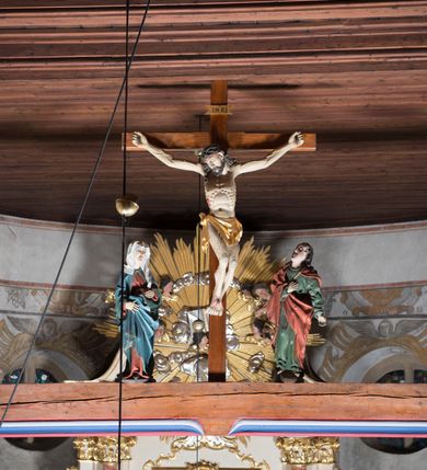 Zdjęcie nr 2: Rzeźbiarska grupa Ukrzyżowania z krucyfiksem pośrodku i stojącymi po bokach figurami: po lewej Matki Boskiej i po prawej św. Jana Ewangelisty. Chrystus przybity do krzyża trzema gwoździami, w lekkim zwisie o delikatnie esowato wygiętej sylwetce, z głową przechyloną na prawe ramię, w koronie cierniowej na głowie. Nogi ugięte, stopy w układzie prawa na lewą. Ciało głęboko rzeźbione o naturalistycznie potraktowanej anatomii podkreślającej żebra i żyłkowanie na nogach i rękach z zaznaczonymi śladami męki. Twarz szczupła o wyraźnych rysach, zamkniętych oczach, długim nosie i delikatnie rozchylonych ustach; okolona brodą. Włosy długie, w postaci loków opadają na prawe ramię i plecy. Perizonium w formie krótkiej tkaniny, odsłaniającej prawe biodro, ze zwisem wzdłuż prawego boku. Nad głową Chrystusa zawieszony jest titulus w formie poziomej banderoli z napisem „I. N. R. I”. Rzeźba ustawiona jest na półkolistym, brązowym cokole. Krzyż w kolorze naturalnego drewna, titulus i perizonium złocone. Po lewej stronie grupy Ukrzyżowania ustawiona jest pełnoplastyczna figura Matki Boskiej zwróconej trzy czwarte w lewą stronę o delikatnie esowato wygiętej sylwetce z rękami uniesionymi na wysokości bioder. Twarz szczupła o wyraźnych rysach, dużych oczach, spiczastym nosie i ustach ukazanych w wyrazie bólu. Ubrana jest w czerwoną suknię, przewiązaną w talii brązowym paskiem, niebieski płaszcz z zieloną podszewką i srebrną lamówką oraz biały welon na głowie; na stopach ma założone brązowe buty. Draperia szat jest głęboka. Po prawej stronie grupy stoi figura św. Jana Ewangelisty, zwróconego trzy czwarte w prawo, z głową uniesioną do góry, z prawą ręką złożoną na piersi, a lewą odsuniętą w bok. Twarz podłużna, z małymi ustami, długim nosem i oczami wzniesionymi do góry, okolona długimi i brązowymi włosami, opadającymi lokami na plecy i ramiona. Święty ubrany jest w zieloną suknię z długimi rękawami oraz czerwony płaszcz. Rzeźba ustawiona jest na półkolistym, brązowym cokole. 