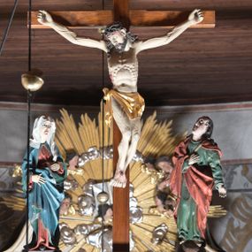 Zdjęcie nr 1: Rzeźbiarska grupa Ukrzyżowania z krucyfiksem pośrodku i stojącymi po bokach figurami: po lewej Matki Boskiej i po prawej św. Jana Ewangelisty. Chrystus przybity do krzyża trzema gwoździami, w lekkim zwisie o delikatnie esowato wygiętej sylwetce, z głową przechyloną na prawe ramię, w koronie cierniowej na głowie. Nogi ugięte, stopy w układzie prawa na lewą. Ciało głęboko rzeźbione o naturalistycznie potraktowanej anatomii podkreślającej żebra i żyłkowanie na nogach i rękach z zaznaczonymi śladami męki. Twarz szczupła o wyraźnych rysach, zamkniętych oczach, długim nosie i delikatnie rozchylonych ustach; okolona brodą. Włosy długie, w postaci loków opadają na prawe ramię i plecy. Perizonium w formie krótkiej tkaniny, odsłaniającej prawe biodro, ze zwisem wzdłuż prawego boku. Nad głową Chrystusa zawieszony jest titulus w formie poziomej banderoli z napisem „I. N. R. I”. Rzeźba ustawiona jest na półkolistym, brązowym cokole. Krzyż w kolorze naturalnego drewna, titulus i perizonium złocone. Po lewej stronie grupy Ukrzyżowania ustawiona jest pełnoplastyczna figura Matki Boskiej zwróconej trzy czwarte w lewą stronę o delikatnie esowato wygiętej sylwetce z rękami uniesionymi na wysokości bioder. Twarz szczupła o wyraźnych rysach, dużych oczach, spiczastym nosie i ustach ukazanych w wyrazie bólu. Ubrana jest w czerwoną suknię, przewiązaną w talii brązowym paskiem, niebieski płaszcz z zieloną podszewką i srebrną lamówką oraz biały welon na głowie; na stopach ma założone brązowe buty. Draperia szat jest głęboka. Po prawej stronie grupy stoi figura św. Jana Ewangelisty, zwróconego trzy czwarte w prawo, z głową uniesioną do góry, z prawą ręką złożoną na piersi, a lewą odsuniętą w bok. Twarz podłużna, z małymi ustami, długim nosem i oczami wzniesionymi do góry, okolona długimi i brązowymi włosami, opadającymi lokami na plecy i ramiona. Święty ubrany jest w zieloną suknię z długimi rękawami oraz czerwony płaszcz. Rzeźba ustawiona jest na półkolistym, brązowym cokole. 