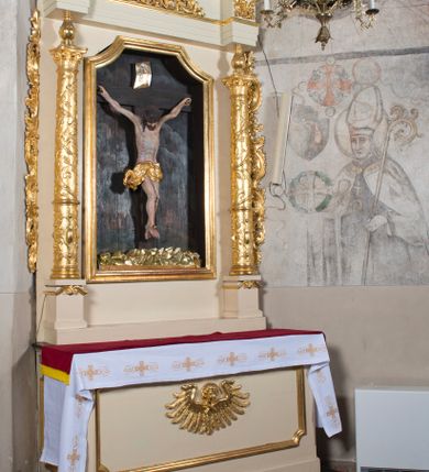 Zdjęcie nr 2: Ołtarz przyścienny, architektoniczny, jednoosiowy, jednokondygnacyjny. Ołtarz prostopadłościenny, skrzyniowy, mensa drewniana z kamiennym portatylem. Antepedium zdobione prostokątną płyciną o wklęsłych narożach, w której rzeźbiona uskrzydlona główka anielska. Retabulum ujęte parą kolumn, ustawionych na bazach. Trzony kolumn dekorowane w jednej trzeciej wysokości ornamentem okuciowym z rautami i kaboszonami, powyżej przewiązane opaską i zdobione w górnej części płaskorzeźbioną wicią winorośli. Powyżej kapiteli ustawione wazony wspierające wyłamane ponad nimi belkowanie z  trójkątnym, przerwanym przyczółkiem.  Pośrodku zwieńczenia pełnoplastyczna figura uskrzydlonego putta stojąca na kartuszu. Nastawa po bokach dekorowana ażurowymi, wąskimi uszakami formowanymi ze splotów z akantu i ornamentu rocaille&#039;owego. Fryz zdobiony rzeźbioną wicią roślinną z rozetami. Pole środkowe w formie prostokątnej wnęki o wklęsłych górnych narożach, ujętej profilowaną ramą; wewnątrz rzeźbiony krucyfiks na malowanym tle Jerozolimy. Całość struktury ołtarza polichromowana w odcieniu jasnego beżu, detal architektoniczny, ornamentyka i profilowania złocone; pole kartusza i szata aniołka srebrzone.

