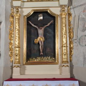 Zdjęcie nr 1: Ołtarz przyścienny, architektoniczny, jednoosiowy, jednokondygnacyjny. Ołtarz prostopadłościenny, skrzyniowy, mensa drewniana z kamiennym portatylem. Antepedium zdobione prostokątną płyciną o wklęsłych narożach, w której rzeźbiona uskrzydlona główka anielska. Retabulum ujęte parą kolumn, ustawionych na bazach. Trzony kolumn dekorowane w jednej trzeciej wysokości ornamentem okuciowym z rautami i kaboszonami, powyżej przewiązane opaską i zdobione w górnej części płaskorzeźbioną wicią winorośli. Powyżej kapiteli ustawione wazony wspierające wyłamane ponad nimi belkowanie z  trójkątnym, przerwanym przyczółkiem.  Pośrodku zwieńczenia pełnoplastyczna figura uskrzydlonego putta stojąca na kartuszu. Nastawa po bokach dekorowana ażurowymi, wąskimi uszakami formowanymi ze splotów z akantu i ornamentu rocaille&#039;owego. Fryz zdobiony rzeźbioną wicią roślinną z rozetami. Pole środkowe w formie prostokątnej wnęki o wklęsłych górnych narożach, ujętej profilowaną ramą; wewnątrz rzeźbiony krucyfiks na malowanym tle Jerozolimy. Całość struktury ołtarza polichromowana w odcieniu jasnego beżu, detal architektoniczny, ornamentyka i profilowania złocone; pole kartusza i szata aniołka srebrzone.

