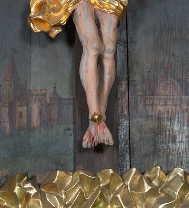 Zdjęcie nr 4: Krucyfiks umieszczony w polu środkowym ołtarza na tle malowanego pejzażu Jerozolimy, u dołu płaskorzeźbiona i złocona skała. Figura Chrystusa pełnoplastyczna ścięta w partii bioder, przybita do drewnianego krzyża trzema gwoździami. Sylwetka wychudzona z wyraźnie zaznaczoną muskulaturą i układem kostnym zwłaszcza w partii klatki piersiowej. Ramiona uniesione wysoko tworzą głęboki łuk. Ręce lekko zgięte, palce dłoni zaciśnięte. Głowa w koronie cierniowej opada na klatkę piersiową, nieznacznie przechylona w prawą stronę. Nogi lekko ugięte, stopy skrzyżowane, przybite jednym gwoździem. Twarz Chrystusa pociągła, okolona długimi włosami opadającymi na prawe ramię oraz krótką brodą. Oczy zamknięte, nos prosty, usta wąskie i rozchylone. Wokół bioder Chrystus ma zawieszone krótkie perizonium, bogato drapowane, z rozwianą kaskadą fałd przy prawym boku. Nad głową wisi titulus w formie banderoli z napisem „IN/RI”.Karnacja ciała w odcieniu szaro-różowym z zaznaczonymi śladami męki  na całym ciele. Włosy i zarost ciemnobrązowe, perizonium i gwoździe złocone, titulus złocony i srebrzony.

