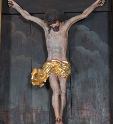 Zdjęcie nr 1: Krucyfiks umieszczony w polu środkowym ołtarza na tle malowanego pejzażu Jerozolimy, u dołu płaskorzeźbiona i złocona skała. Figura Chrystusa pełnoplastyczna ścięta w partii bioder, przybita do drewnianego krzyża trzema gwoździami. Sylwetka wychudzona z wyraźnie zaznaczoną muskulaturą i układem kostnym zwłaszcza w partii klatki piersiowej. Ramiona uniesione wysoko tworzą głęboki łuk. Ręce lekko zgięte, palce dłoni zaciśnięte. Głowa w koronie cierniowej opada na klatkę piersiową, nieznacznie przechylona w prawą stronę. Nogi lekko ugięte, stopy skrzyżowane, przybite jednym gwoździem. Twarz Chrystusa pociągła, okolona długimi włosami opadającymi na prawe ramię oraz krótką brodą. Oczy zamknięte, nos prosty, usta wąskie i rozchylone. Wokół bioder Chrystus ma zawieszone krótkie perizonium, bogato drapowane, z rozwianą kaskadą fałd przy prawym boku. Nad głową wisi titulus w formie banderoli z napisem „IN/RI”.Karnacja ciała w odcieniu szaro-różowym z zaznaczonymi śladami męki  na całym ciele. Włosy i zarost ciemnobrązowe, perizonium i gwoździe złocone, titulus złocony i srebrzony.

