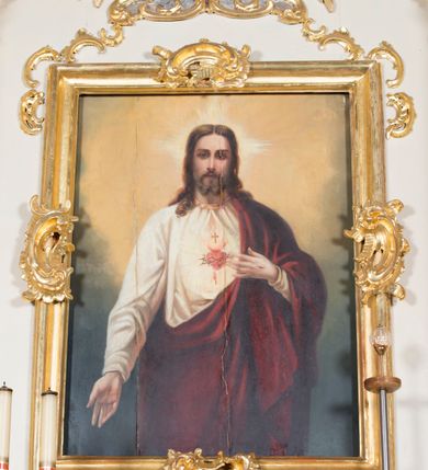 Zdjęcie nr 1: Obraz w kształcie stojącego prostokąta. W centrum kompozycji ukazany Chrystus w półpostaci, frontalnie, z lewą ręką wskazującą na gorejące na piersi serce, prawą spuszczoną w dół. Ubrany w długą, białą suknię z długimi rękawami, u szyi obwiedzioną złotą lamówką oraz czerwony płaszcz, założony na lewe ramię. Na piersi Chrystusa gorejące serce na tle promienistej glorii, z zatkniętym u szczytu małym krzyżykiem, oplecione koroną cierniową. Twarz podłużna z długim i wąskim nosem, małymi oczami, ze wzrokiem skierowanym na wprost, okolona krótką brodą, rozdzieloną na dwa pukle oraz długimi włosami, z przedziałkiem pośrodku, spływającymi długimi puklami na ramiona i plecy. Wokół głowy Chrystusa nimb krzyżowy.
