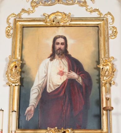 Zdjęcie nr 1: Obraz w kształcie stojącego prostokąta. W centrum kompozycji ukazany Chrystus w półpostaci, frontalnie, z lewą ręką wskazującą na gorejące na piersi serce, prawą spuszczoną w dół. Ubrany w długą, białą suknię z długimi rękawami, u szyi obwiedzioną złotą lamówką oraz czerwony płaszcz, założony na lewe ramię. Na piersi Chrystusa gorejące serce na tle promienistej glorii, z zatkniętym u szczytu małym krzyżykiem, oplecione koroną cierniową. Twarz podłużna z długim i wąskim nosem, małymi oczami, ze wzrokiem skierowanym na wprost, okolona krótką brodą, rozdzieloną na dwa pukle oraz długimi włosami, z przedziałkiem pośrodku, spływającymi długimi puklami na ramiona i plecy. Wokół głowy Chrystusa nimb krzyżowy.
