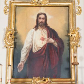 Zdjęcie nr 1: Obraz w kształcie stojącego prostokąta. W centrum kompozycji ukazany Chrystus w półpostaci, frontalnie, z lewą ręką wskazującą na gorejące na piersi serce, prawą spuszczoną w dół. Ubrany w długą, białą suknię z długimi rękawami, u szyi obwiedzioną złotą lamówką oraz czerwony płaszcz, założony na lewe ramię. Na piersi Chrystusa gorejące serce na tle promienistej glorii, z zatkniętym u szczytu małym krzyżykiem, oplecione koroną cierniową. Twarz podłużna z długim i wąskim nosem, małymi oczami, ze wzrokiem skierowanym na wprost, okolona krótką brodą, rozdzieloną na dwa pukle oraz długimi włosami, z przedziałkiem pośrodku, spływającymi długimi puklami na ramiona i plecy. Wokół głowy Chrystusa nimb krzyżowy.
