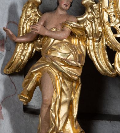 Zdjęcie nr 2: Rzeźba ścięta z tyłu. Figura ustawiona w kontrapoście z prawą nogą ugiętą w kolanie, o esowatym układzie ciała, z głową skierowaną w lewą stronę i obiema rękami wyciągniętymi w prawo. Twarz młodzieńcza, pełna, o dużych oczach, małym nosie i delikatnie rozchylonych ustach. Włosy w postaci grubych pasm opadają na tył głowy. Anioł ubrany jest w szatę przepasaną diagonalnie przez tors, ściśle przylegającą do ciała i odsłaniającą prawą nogę oraz płaszcz przerzucony przez prawe ramię i spoczywający na linii bioder. Skrzydła rozpostarte na boki. Polichromia naturalistyczna w partiach ciała, szaty i skrzydła złocone.