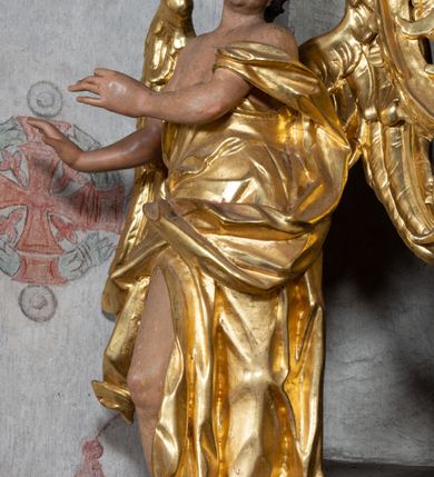 Zdjęcie nr 4: Rzeźba ścięta z tyłu. Figura ustawiona w kontrapoście z prawą nogą ugiętą w kolanie, o esowatym układzie ciała, z głową skierowaną w lewą stronę i obiema rękami wyciągniętymi w prawo. Twarz młodzieńcza, pełna, o dużych oczach, małym nosie i delikatnie rozchylonych ustach. Włosy w postaci grubych pasm opadają na tył głowy. Anioł ubrany jest w szatę przepasaną diagonalnie przez tors, ściśle przylegającą do ciała i odsłaniającą prawą nogę oraz płaszcz przerzucony przez prawe ramię i spoczywający na linii bioder. Skrzydła rozpostarte na boki. Polichromia naturalistyczna w partiach ciała, szaty i skrzydła złocone.