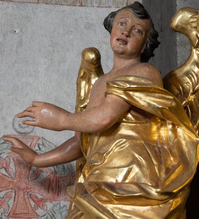 Zdjęcie nr 5: Rzeźba ścięta z tyłu. Figura ustawiona w kontrapoście z prawą nogą ugiętą w kolanie, o esowatym układzie ciała, z głową skierowaną w lewą stronę i obiema rękami wyciągniętymi w prawo. Twarz młodzieńcza, pełna, o dużych oczach, małym nosie i delikatnie rozchylonych ustach. Włosy w postaci grubych pasm opadają na tył głowy. Anioł ubrany jest w szatę przepasaną diagonalnie przez tors, ściśle przylegającą do ciała i odsłaniającą prawą nogę oraz płaszcz przerzucony przez prawe ramię i spoczywający na linii bioder. Skrzydła rozpostarte na boki. Polichromia naturalistyczna w partiach ciała, szaty i skrzydła złocone.