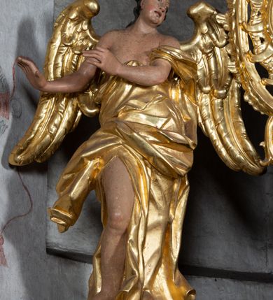 Zdjęcie nr 3: Rzeźba ścięta z tyłu. Figura ustawiona w kontrapoście z prawą nogą ugiętą w kolanie, o esowatym układzie ciała, z głową skierowaną w lewą stronę i obiema rękami wyciągniętymi w prawo. Twarz młodzieńcza, pełna, o dużych oczach, małym nosie i delikatnie rozchylonych ustach. Włosy w postaci grubych pasm opadają na tył głowy. Anioł ubrany jest w szatę przepasaną diagonalnie przez tors, ściśle przylegającą do ciała i odsłaniającą prawą nogę oraz płaszcz przerzucony przez prawe ramię i spoczywający na linii bioder. Skrzydła rozpostarte na boki. Polichromia naturalistyczna w partiach ciała, szaty i skrzydła złocone.