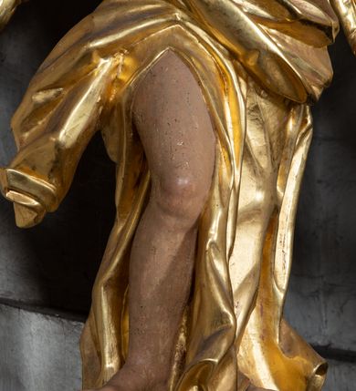 Zdjęcie nr 8: Rzeźba ścięta z tyłu. Figura ustawiona w kontrapoście z prawą nogą ugiętą w kolanie, o esowatym układzie ciała, z głową skierowaną w lewą stronę i obiema rękami wyciągniętymi w prawo. Twarz młodzieńcza, pełna, o dużych oczach, małym nosie i delikatnie rozchylonych ustach. Włosy w postaci grubych pasm opadają na tył głowy. Anioł ubrany jest w szatę przepasaną diagonalnie przez tors, ściśle przylegającą do ciała i odsłaniającą prawą nogę oraz płaszcz przerzucony przez prawe ramię i spoczywający na linii bioder. Skrzydła rozpostarte na boki. Polichromia naturalistyczna w partiach ciała, szaty i skrzydła złocone.