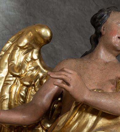 Zdjęcie nr 6: Rzeźba ścięta z tyłu. Figura ustawiona w kontrapoście z prawą nogą ugiętą w kolanie, o esowatym układzie ciała, z głową skierowaną w lewą stronę i obiema rękami wyciągniętymi w prawo. Twarz młodzieńcza, pełna, o dużych oczach, małym nosie i delikatnie rozchylonych ustach. Włosy w postaci grubych pasm opadają na tył głowy. Anioł ubrany jest w szatę przepasaną diagonalnie przez tors, ściśle przylegającą do ciała i odsłaniającą prawą nogę oraz płaszcz przerzucony przez prawe ramię i spoczywający na linii bioder. Skrzydła rozpostarte na boki. Polichromia naturalistyczna w partiach ciała, szaty i skrzydła złocone.