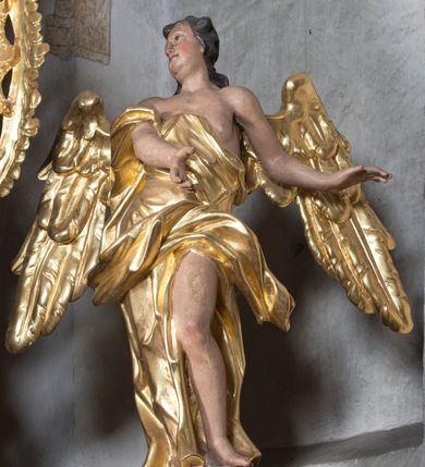 Zdjęcie nr 2: Rzeźba ścięta z tyłu. Figura ustawiona w kontrapoście o esowatym układzie ciała, z głową skierowaną w prawą stronę i obiema rękami wyciągniętymi w lewą stronę. Twarz młodzieńcza, pełna, o dużych oczach, małym nosie i delikatnie rozchylonych ustach. Włosy krótkie, w postaci grubych pasm opadających na tył głowy. Anioł ubrany jest w szatę przełożoną diagonalnie przez tors, ściśle przylegającą do ciała i odsłaniającą lewą nogę oraz płaszcz przerzucony przez prawe ramię i spoczywający na linii bioder. Skrzydła rozpostarte na boki. Polichromia naturalistyczna w partiach ciała, szaty i skrzydła złocone.