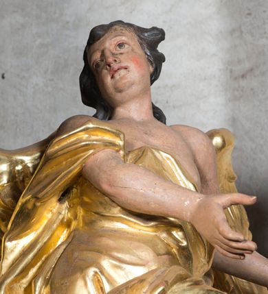 Zdjęcie nr 9: Rzeźba ścięta z tyłu. Figura ustawiona w kontrapoście o esowatym układzie ciała, z głową skierowaną w prawą stronę i obiema rękami wyciągniętymi w lewą stronę. Twarz młodzieńcza, pełna, o dużych oczach, małym nosie i delikatnie rozchylonych ustach. Włosy krótkie, w postaci grubych pasm opadających na tył głowy. Anioł ubrany jest w szatę przełożoną diagonalnie przez tors, ściśle przylegającą do ciała i odsłaniającą lewą nogę oraz płaszcz przerzucony przez prawe ramię i spoczywający na linii bioder. Skrzydła rozpostarte na boki. Polichromia naturalistyczna w partiach ciała, szaty i skrzydła złocone.