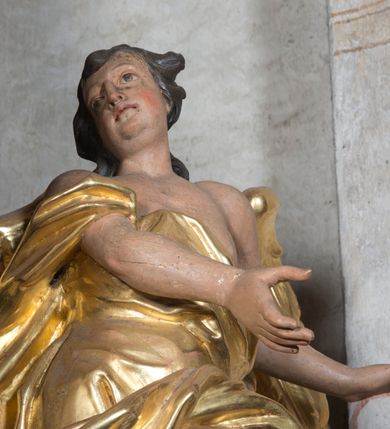 Zdjęcie nr 6: Rzeźba ścięta z tyłu. Figura ustawiona w kontrapoście o esowatym układzie ciała, z głową skierowaną w prawą stronę i obiema rękami wyciągniętymi w lewą stronę. Twarz młodzieńcza, pełna, o dużych oczach, małym nosie i delikatnie rozchylonych ustach. Włosy krótkie, w postaci grubych pasm opadających na tył głowy. Anioł ubrany jest w szatę przełożoną diagonalnie przez tors, ściśle przylegającą do ciała i odsłaniającą lewą nogę oraz płaszcz przerzucony przez prawe ramię i spoczywający na linii bioder. Skrzydła rozpostarte na boki. Polichromia naturalistyczna w partiach ciała, szaty i skrzydła złocone.
