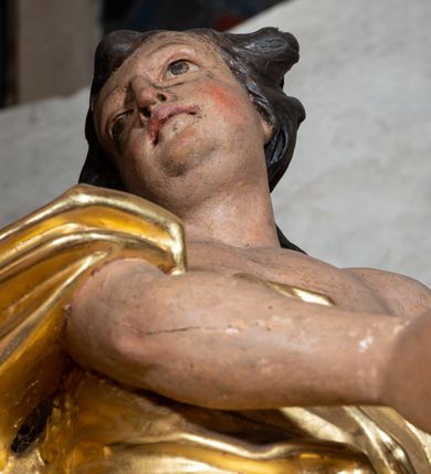 Zdjęcie nr 10: Rzeźba ścięta z tyłu. Figura ustawiona w kontrapoście o esowatym układzie ciała, z głową skierowaną w prawą stronę i obiema rękami wyciągniętymi w lewą stronę. Twarz młodzieńcza, pełna, o dużych oczach, małym nosie i delikatnie rozchylonych ustach. Włosy krótkie, w postaci grubych pasm opadających na tył głowy. Anioł ubrany jest w szatę przełożoną diagonalnie przez tors, ściśle przylegającą do ciała i odsłaniającą lewą nogę oraz płaszcz przerzucony przez prawe ramię i spoczywający na linii bioder. Skrzydła rozpostarte na boki. Polichromia naturalistyczna w partiach ciała, szaty i skrzydła złocone.