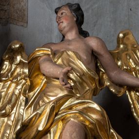 Zdjęcie nr 1: Rzeźba ścięta z tyłu. Figura ustawiona w kontrapoście o esowatym układzie ciała, z głową skierowaną w prawą stronę i obiema rękami wyciągniętymi w lewą stronę. Twarz młodzieńcza, pełna, o dużych oczach, małym nosie i delikatnie rozchylonych ustach. Włosy krótkie, w postaci grubych pasm opadających na tył głowy. Anioł ubrany jest w szatę przełożoną diagonalnie przez tors, ściśle przylegającą do ciała i odsłaniającą lewą nogę oraz płaszcz przerzucony przez prawe ramię i spoczywający na linii bioder. Skrzydła rozpostarte na boki. Polichromia naturalistyczna w partiach ciała, szaty i skrzydła złocone.