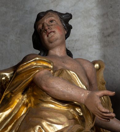 Zdjęcie nr 8: Rzeźba ścięta z tyłu. Figura ustawiona w kontrapoście o esowatym układzie ciała, z głową skierowaną w prawą stronę i obiema rękami wyciągniętymi w lewą stronę. Twarz młodzieńcza, pełna, o dużych oczach, małym nosie i delikatnie rozchylonych ustach. Włosy krótkie, w postaci grubych pasm opadających na tył głowy. Anioł ubrany jest w szatę przełożoną diagonalnie przez tors, ściśle przylegającą do ciała i odsłaniającą lewą nogę oraz płaszcz przerzucony przez prawe ramię i spoczywający na linii bioder. Skrzydła rozpostarte na boki. Polichromia naturalistyczna w partiach ciała, szaty i skrzydła złocone.