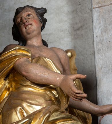 Zdjęcie nr 7: Rzeźba ścięta z tyłu. Figura ustawiona w kontrapoście o esowatym układzie ciała, z głową skierowaną w prawą stronę i obiema rękami wyciągniętymi w lewą stronę. Twarz młodzieńcza, pełna, o dużych oczach, małym nosie i delikatnie rozchylonych ustach. Włosy krótkie, w postaci grubych pasm opadających na tył głowy. Anioł ubrany jest w szatę przełożoną diagonalnie przez tors, ściśle przylegającą do ciała i odsłaniającą lewą nogę oraz płaszcz przerzucony przez prawe ramię i spoczywający na linii bioder. Skrzydła rozpostarte na boki. Polichromia naturalistyczna w partiach ciała, szaty i skrzydła złocone.