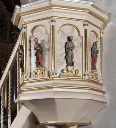 Zdjęcie nr 2: Ambona przyścienna, wisząca, z dostępem schodkami z lewej strony. Podwieszenie ośmioboczne, zwężające się ku dołowi, zakończone trzema spiętymi kabłąkami. Kosz na planie ośmioboku ze ściankami oddzielonymi półkolumnami, pomiędzy którymi płyciny w formie stojącego prostokąta zamkniętego łukiem nadwieszonym. W polach płycin, na cokołach ustawione figury ewangelistów i Chrystusa Salwatora. Od lewej: św. Jan, św. Marek, św. Łukasz i Chrystus Salwator. Ewangeliści z księgami w dłoniach, Chrystus z jabłkiem królewskim. Ubrani są w długie suknie i płaszcze przewieszone przez jedno ramię. Parapet w formie niepełnego belkowania z wyłamanym gzymsem Schody jednobiegowe, ośmiostopniowe z balustradą o tralkach w kształcie skręconych kolumn. Ambona polichromowana na beżowo, ze złoconymi i srebrzonymi detalami. Tło płycin zdobione imitacją marmoryzacji.