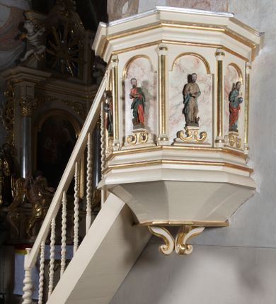 Zdjęcie nr 1: Ambona przyścienna, wisząca, z dostępem schodkami z lewej strony. Podwieszenie ośmioboczne, zwężające się ku dołowi, zakończone trzema spiętymi kabłąkami. Kosz na planie ośmioboku ze ściankami oddzielonymi półkolumnami, pomiędzy którymi płyciny w formie stojącego prostokąta zamkniętego łukiem nadwieszonym. W polach płycin, na cokołach ustawione figury ewangelistów i Chrystusa Salwatora. Od lewej: św. Jan, św. Marek, św. Łukasz i Chrystus Salwator. Ewangeliści z księgami w dłoniach, Chrystus z jabłkiem królewskim. Ubrani są w długie suknie i płaszcze przewieszone przez jedno ramię. Parapet w formie niepełnego belkowania z wyłamanym gzymsem Schody jednobiegowe, ośmiostopniowe z balustradą o tralkach w kształcie skręconych kolumn. Ambona polichromowana na beżowo, ze złoconymi i srebrzonymi detalami. Tło płycin zdobione imitacją marmoryzacji.