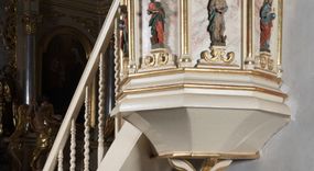 Zdjęcie nr 1: Ambona przyścienna, wisząca, z dostępem schodkami z lewej strony. Podwieszenie ośmioboczne, zwężające się ku dołowi, zakończone trzema spiętymi kabłąkami. Kosz na planie ośmioboku ze ściankami oddzielonymi półkolumnami, pomiędzy którymi płyciny w formie stojącego prostokąta zamkniętego łukiem nadwieszonym. W polach płycin, na cokołach ustawione figury ewangelistów i Chrystusa Salwatora. Od lewej: św. Jan, św. Marek, św. Łukasz i Chrystus Salwator. Ewangeliści z księgami w dłoniach, Chrystus z jabłkiem królewskim. Ubrani są w długie suknie i płaszcze przewieszone przez jedno ramię. Parapet w formie niepełnego belkowania z wyłamanym gzymsem Schody jednobiegowe, ośmiostopniowe z balustradą o tralkach w kształcie skręconych kolumn. Ambona polichromowana na beżowo, ze złoconymi i srebrzonymi detalami. Tło płycin zdobione imitacją marmoryzacji.