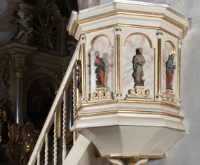 Zdjęcie nr 1: Ambona przyścienna, wisząca, z dostępem schodkami z lewej strony. Podwieszenie ośmioboczne, zwężające się ku dołowi, zakończone trzema spiętymi kabłąkami. Kosz na planie ośmioboku ze ściankami oddzielonymi półkolumnami, pomiędzy którymi płyciny w formie stojącego prostokąta zamkniętego łukiem nadwieszonym. W polach płycin, na cokołach ustawione figury ewangelistów i Chrystusa Salwatora. Od lewej: św. Jan, św. Marek, św. Łukasz i Chrystus Salwator. Ewangeliści z księgami w dłoniach, Chrystus z jabłkiem królewskim. Ubrani są w długie suknie i płaszcze przewieszone przez jedno ramię. Parapet w formie niepełnego belkowania z wyłamanym gzymsem Schody jednobiegowe, ośmiostopniowe z balustradą o tralkach w kształcie skręconych kolumn. Ambona polichromowana na beżowo, ze złoconymi i srebrzonymi detalami. Tło płycin zdobione imitacją marmoryzacji.