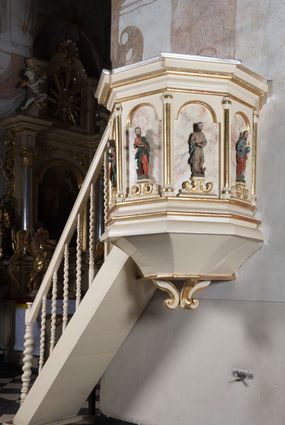 Zdjęcie nr 1: Ambona przyścienna, wisząca, z dostępem schodkami z lewej strony. Podwieszenie ośmioboczne, zwężające się ku dołowi, zakończone trzema spiętymi kabłąkami. Kosz na planie ośmioboku ze ściankami oddzielonymi półkolumnami, pomiędzy którymi płyciny w formie stojącego prostokąta zamkniętego łukiem nadwieszonym. W polach płycin, na cokołach ustawione figury ewangelistów i Chrystusa Salwatora. Od lewej: św. Jan, św. Marek, św. Łukasz i Chrystus Salwator. Ewangeliści z księgami w dłoniach, Chrystus z jabłkiem królewskim. Ubrani są w długie suknie i płaszcze przewieszone przez jedno ramię. Parapet w formie niepełnego belkowania z wyłamanym gzymsem Schody jednobiegowe, ośmiostopniowe z balustradą o tralkach w kształcie skręconych kolumn. Ambona polichromowana na beżowo, ze złoconymi i srebrzonymi detalami. Tło płycin zdobione imitacją marmoryzacji.