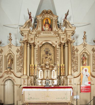 Zdjęcie nr 1: Ołtarz architektoniczny, jednokondygnacyjny, trójosiowy, ze zwieńczeniem. Ołtarz prostopadłościenny, z drewnianą mensą. Antepedium w formie haftowanej tkaniny zdobionej po środku motywem zwieńczonego krzyżem monogramu maryjnego z koroną, ujętego dwunastoma gwiazdami, natomiast w dolnych narożach gałązkami z kwiatami białych lilii.  Tabernakulum z tronem wystawienia w formie jednokondygnacyjnej, trójosiowej bryły architektonicznej, osadzonej na wysokim cokole. Tabernakulum wbudowane w osi środkowej cokołu, drzwiczki prostokątne, zdobione płaskorzeźbionym motywem kielicha eucharystycznego. Flankowane po skrajnych bokach parą pilastrów osie boczne całej struktury są wklęsłe i cofnięte względem osi środkowej ujętej parą kolumienek. Podpory wspierają wyłamujące się ponad nimi belkowanie z gzymsem po środku w formie łuku koszowego. Część środkowa w formie niszy zamkniętej motywem konchy. W zwieńczeniu figurka baranka paschalnego ujętego promienistą glorią, ukazanego na owalnej podstawie osadzonej na 4 wolutowych kabłąkach. Po bokach para figur adorujących aniołów. Cokół zdobiony motywami uskrzydlonych główek anielskich, rocaille’i oraz gałązek z kiściami winnej latorośli. Pola osi bocznych wypełnia para podwieszeń w formie gałązek różanych, partie fryzu pojedyncze rozetki. Struktura tabernakulum i tronu wystawienia jest złocona i srebrzona.   Retabulum osadzone na zdwojonym, zryzalitowanym uskokowo po bokach cokole. Część środkowa ujęta dwoma parami kolumn – wewnętrzne ustawione na tle pary pilastrów, zewnętrzne na tle pary filarów. Podpory dźwigają wyłamujące się ponad nimi belkowanie przerwane w partii architrawu i fryzu. Zwieńczenie ujęte parą spływów wolutowych, zamknięte parą stykających się wolut. Po bokach przerwany, wolutowy przyczółek. W dolnych polach osi bocznych retabulum para półkoliście zamkniętych bramek, ujętych prostą arkadą. Pola osi oddzielone parami stykających się wolut zwieńczonych konchą. W górnej strefie para płytkich niszy zamkniętych półkolistymi łukami nadwieszonymi i dodatkowo ujęta prostym, trójkątnie zamkniętym obramieniem. Osie zwieńczone trzema fragmentami gzymsu oddzielonymi parami wklęsłości.  Pola cokołu osi środkowej retabulum wypełnione ornamentyką roślinną. Część środkowa ujęta parą ażurowych, ornamentalnych  uszaków. Po bokach pola para zwisów w postaci gałązek różanych, w górnych narożach para rozetek. Pola fryzu zdobione rozetami. Pole zwieńczenia w górnej partii dekorowane parą ulistnionych gałązek, po bokach parą kampanuli, dołem w narożach para dużych rozet, pośrodku wić roślinna. Na spływach wolutowych para płomienistych wazonów, a na przyczółku para figur aniołów. Nadłucza bramek osi bocznych zdobione ornamentem roślinnym, nisze ujęte parą stylizowanych kandelabrów, wyżej para rombów z rozetami, pośrodku gwiazda. Na gzymsach wieńczących płomieniste wazony. W polu środkowym obraz w kształcie stojącego prostokąta zamkniętego odcinkowym łukiem nadwieszonym z uskokami, z wizerunkiem Madonny z Dzieciątkiem. W zwieńczeniu obraz w kształcie stojącego prostokąta zamkniętego odcinkowym łukiem nadwieszonym z uskokami, z wizerunkiem Chrystusa Króla. Nisze osi bocznych wypełniają figury, po prawej stronie św. Barbary, po lewej nieznanej świętej. Struktura w kolorze beżowym, detal architektoniczny i ornamentyka złocone.     