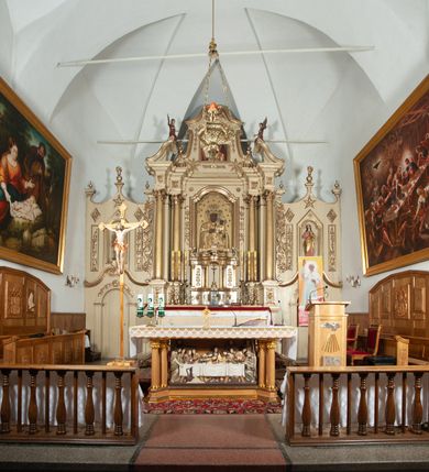 Zdjęcie nr 13: Ołtarz architektoniczny, jednokondygnacyjny, trójosiowy, ze zwieńczeniem. Ołtarz prostopadłościenny, z drewnianą mensą. Antepedium w formie haftowanej tkaniny zdobionej po środku motywem zwieńczonego krzyżem monogramu maryjnego z koroną, ujętego dwunastoma gwiazdami, natomiast w dolnych narożach gałązkami z kwiatami białych lilii.  Tabernakulum z tronem wystawienia w formie jednokondygnacyjnej, trójosiowej bryły architektonicznej, osadzonej na wysokim cokole. Tabernakulum wbudowane w osi środkowej cokołu, drzwiczki prostokątne, zdobione płaskorzeźbionym motywem kielicha eucharystycznego. Flankowane po skrajnych bokach parą pilastrów osie boczne całej struktury są wklęsłe i cofnięte względem osi środkowej ujętej parą kolumienek. Podpory wspierają wyłamujące się ponad nimi belkowanie z gzymsem po środku w formie łuku koszowego. Część środkowa w formie niszy zamkniętej motywem konchy. W zwieńczeniu figurka baranka paschalnego ujętego promienistą glorią, ukazanego na owalnej podstawie osadzonej na 4 wolutowych kabłąkach. Po bokach para figur adorujących aniołów. Cokół zdobiony motywami uskrzydlonych główek anielskich, rocaille’i oraz gałązek z kiściami winnej latorośli. Pola osi bocznych wypełnia para podwieszeń w formie gałązek różanych, partie fryzu pojedyncze rozetki. Struktura tabernakulum i tronu wystawienia jest złocona i srebrzona.   Retabulum osadzone na zdwojonym, zryzalitowanym uskokowo po bokach cokole. Część środkowa ujęta dwoma parami kolumn – wewnętrzne ustawione na tle pary pilastrów, zewnętrzne na tle pary filarów. Podpory dźwigają wyłamujące się ponad nimi belkowanie przerwane w partii architrawu i fryzu. Zwieńczenie ujęte parą spływów wolutowych, zamknięte parą stykających się wolut. Po bokach przerwany, wolutowy przyczółek. W dolnych polach osi bocznych retabulum para półkoliście zamkniętych bramek, ujętych prostą arkadą. Pola osi oddzielone parami stykających się wolut zwieńczonych konchą. W górnej strefie para płytkich niszy zamkniętych półkolistymi łukami nadwieszonymi i dodatkowo ujęta prostym, trójkątnie zamkniętym obramieniem. Osie zwieńczone trzema fragmentami gzymsu oddzielonymi parami wklęsłości.  Pola cokołu osi środkowej retabulum wypełnione ornamentyką roślinną. Część środkowa ujęta parą ażurowych, ornamentalnych  uszaków. Po bokach pola para zwisów w postaci gałązek różanych, w górnych narożach para rozetek. Pola fryzu zdobione rozetami. Pole zwieńczenia w górnej partii dekorowane parą ulistnionych gałązek, po bokach parą kampanuli, dołem w narożach para dużych rozet, pośrodku wić roślinna. Na spływach wolutowych para płomienistych wazonów, a na przyczółku para figur aniołów. Nadłucza bramek osi bocznych zdobione ornamentem roślinnym, nisze ujęte parą stylizowanych kandelabrów, wyżej para rombów z rozetami, pośrodku gwiazda. Na gzymsach wieńczących płomieniste wazony. W polu środkowym obraz w kształcie stojącego prostokąta zamkniętego odcinkowym łukiem nadwieszonym z uskokami, z wizerunkiem Madonny z Dzieciątkiem. W zwieńczeniu obraz w kształcie stojącego prostokąta zamkniętego odcinkowym łukiem nadwieszonym z uskokami, z wizerunkiem Chrystusa Króla. Nisze osi bocznych wypełniają figury, po prawej stronie św. Barbary, po lewej nieznanej świętej. Struktura w kolorze beżowym, detal architektoniczny i ornamentyka złocone.     